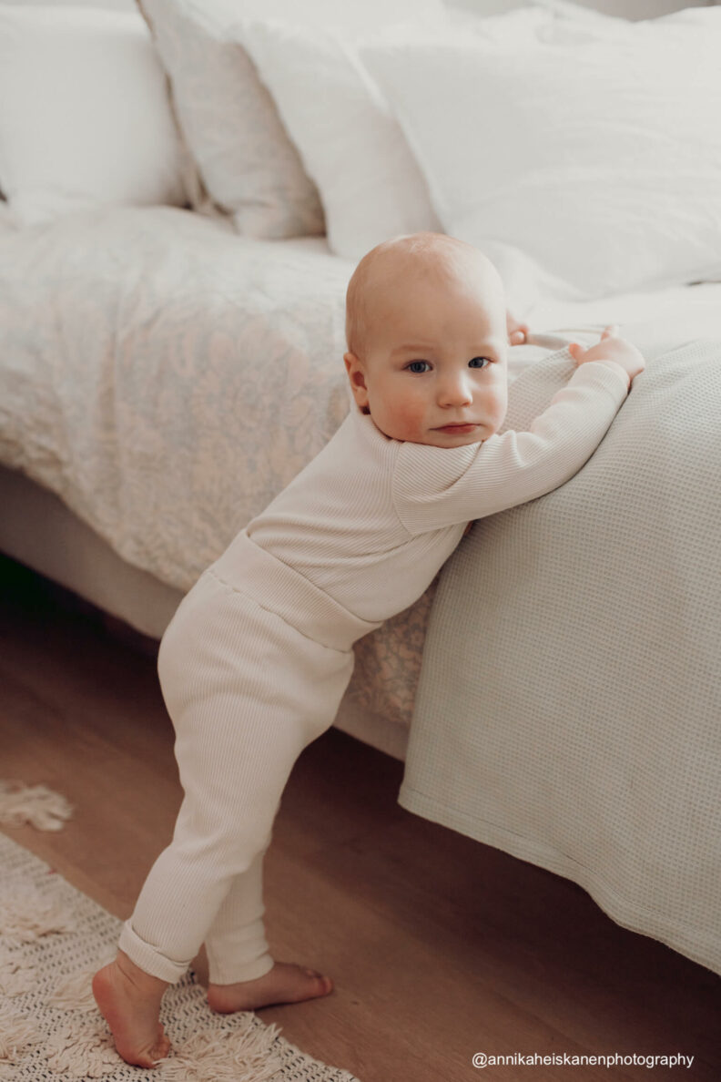 Lapsi seisoo sänkyä vasten valkoisen sävysissä ribbi vaatteissaan