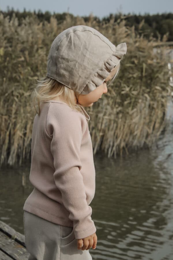 Lapsi kaislikkoisen veden äärellä yllään Uturoosan sävyinen ribbi paita, Pellavan beiget housut ja myssy