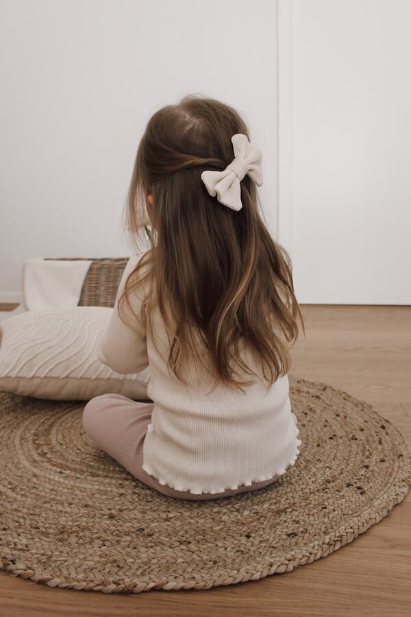 Tyttö istuu lattialla selin kameraan, yllään valkoinen ribbineuloksinen paita ja roosan väriset housut, hiuksissa rusetti.