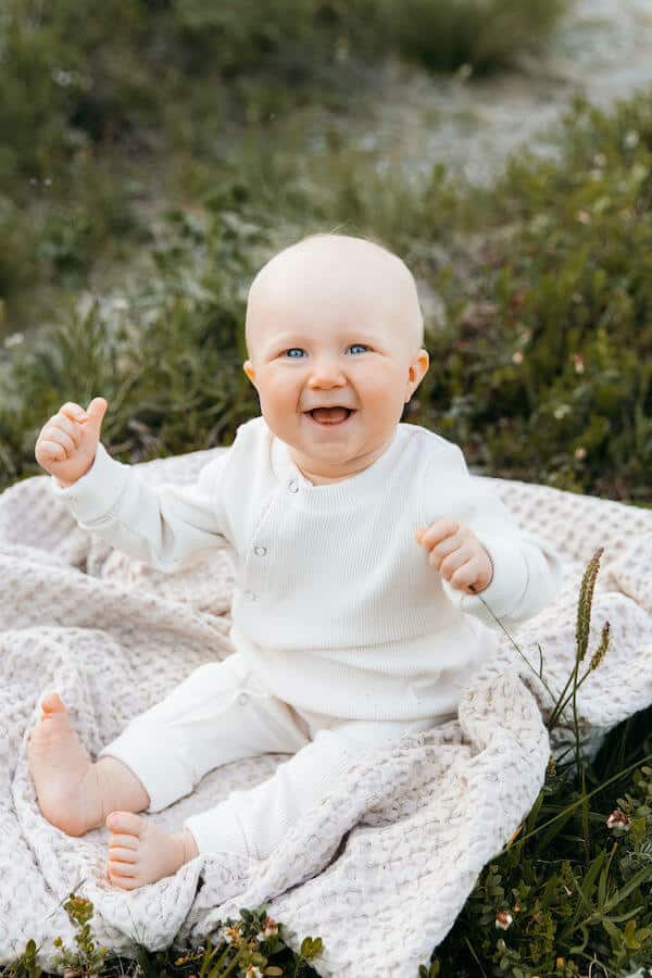 Vauva istuu nurmella peiton päällä valkoisessa ribbihaalarissa ja näyttää peukaloa.